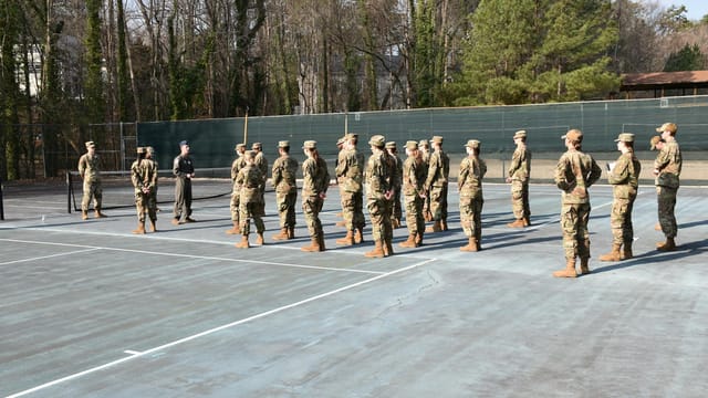 Soldiers in uniform standing in formation on an outdoor tennis court during the day.