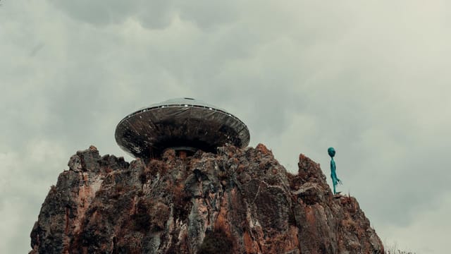 A surreal scene with a UFO and alien standing atop a rugged rock formation against a cloudy sky.
