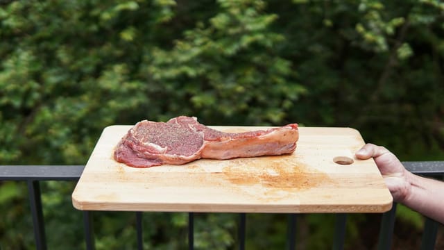 Uncooked seasoned steak on a wooden board with a forest background, ready for cooking.