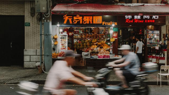 Dynamic image of motorcyclists passing a vibrant fruit stand on Shanghai street.