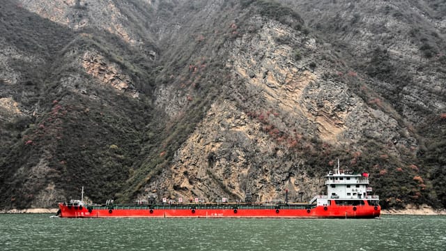 A vibrant red cargo ship sails through a dramatic river gorge surrounded by steep rocky cliffs.