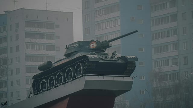 A military tank monument on a platform with residential buildings in the background.