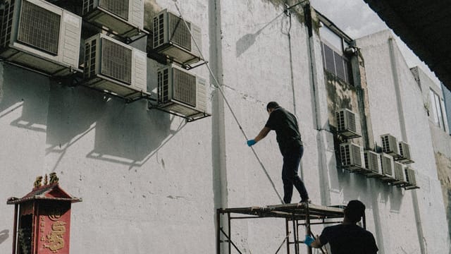 Two workers on scaffold maintain air conditioning units attached to a building, displaying urban upkeep.