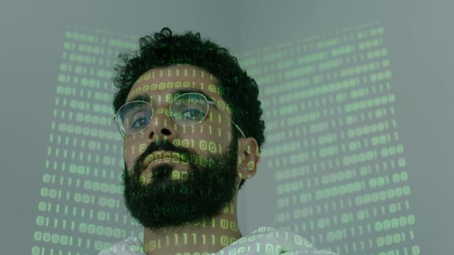 A man with a beard and eyeglasses looking at the camera, illuminated by binary code projection.