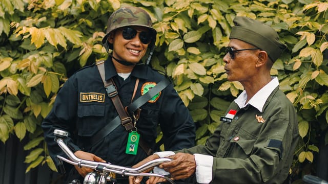 Two men in military-style uniforms conversing next to a bicycle with greenery in the background.