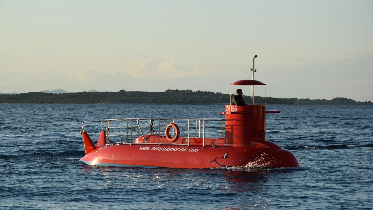 A bright red semi-submarine cruising on a calm ocean with a distant shoreline.