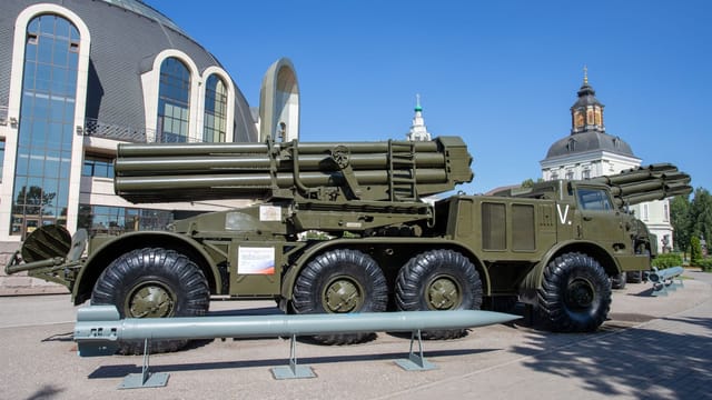 A military missile launcher on exhibit outdoors with a backdrop of unique architecture and clear blue sky.