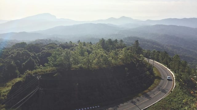 Aerial view of a winding road through lush green forest with misty mountain backdrop in Chiang Mai, Thailand.
