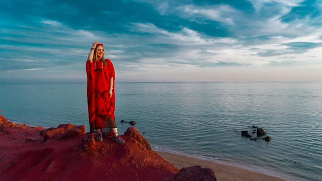 Elegant woman in red dress posing on Hormuz Island's red beach with scenic ocean view.