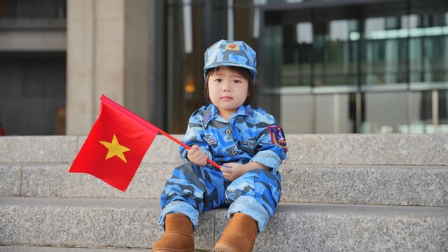 A child dressed in blue camouflage sitting on steps, holding a Vietnamese flag, outdoors.