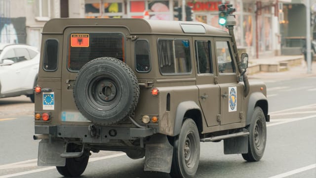 A rugged military vehicle travels through an urban street in Albania, signifying mobility and readiness.