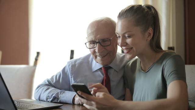 Smiling young woman in casual clothes showing smartphone to interested senior grandfather in formal shirt and eyeglasses while sitting at table near laptop