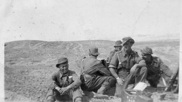 A group of soldiers in a black and white photograph from World War II, outdoors on rugged terrain.
