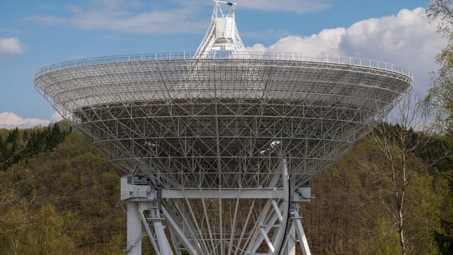 Illustrative image: A large radio telescope set against a backdrop of trees and clouds, capturing celestial signals.