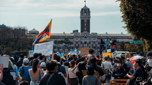Large crowd protesting near government building with flags and signs in urban setting.