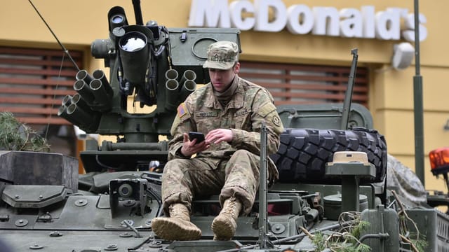 A soldier in camouflage resting on an armored vehicle, using a smartphone outside a McDonald's.