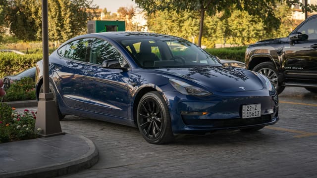 Electric blue Tesla Model 3 parked in sunlit commercial parking area.