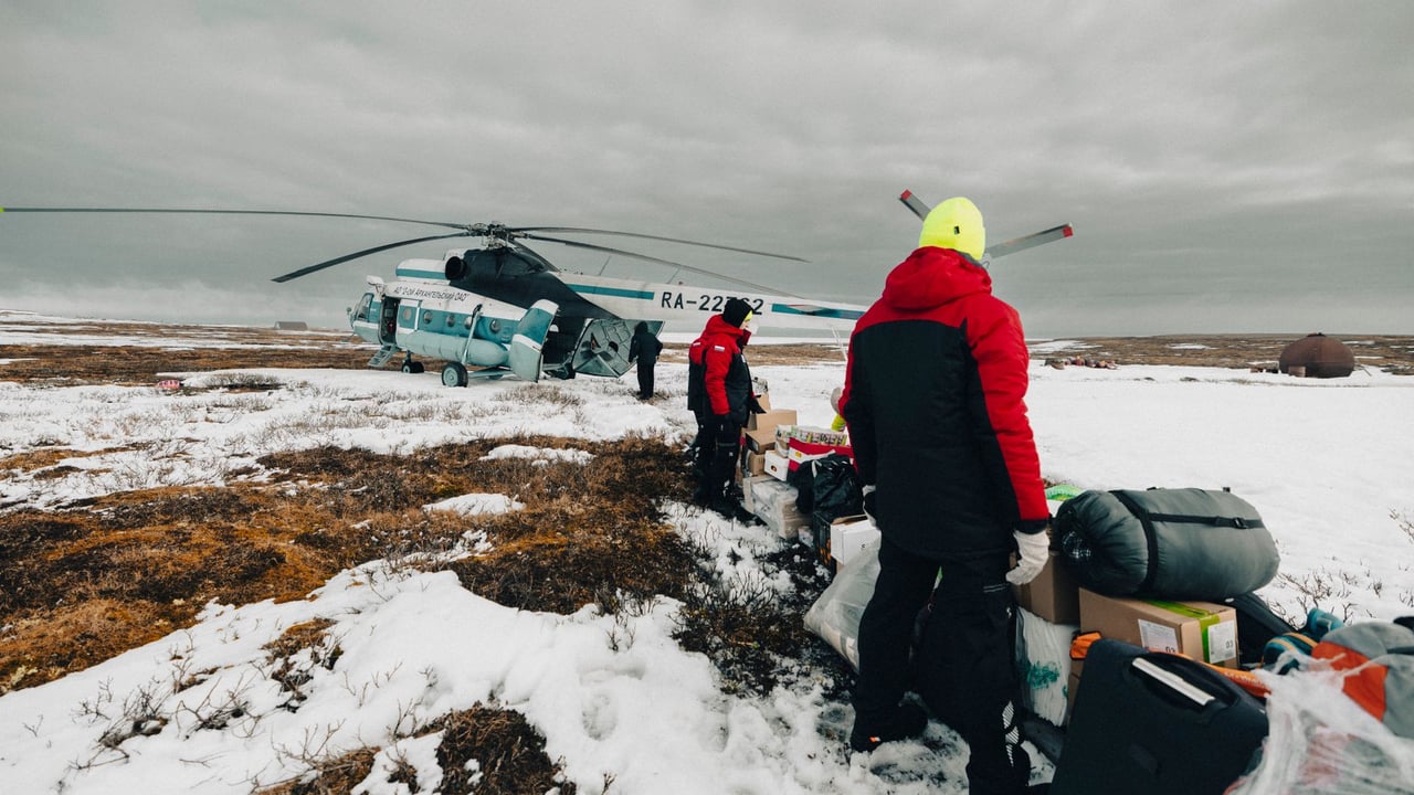Weatherproofed explorers unloading essentials from helicopter in icy tundra landscape.