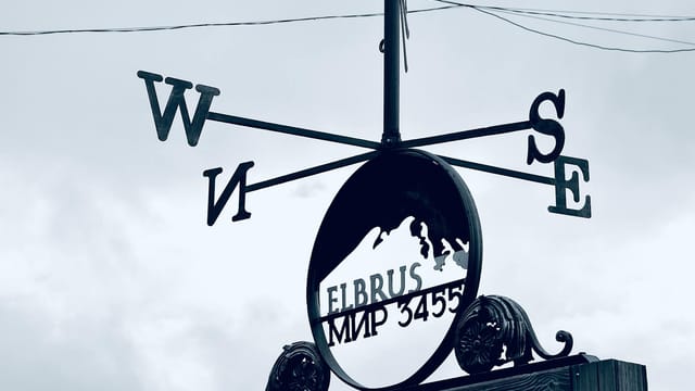 Metal weather vane featuring cardinal directions near Mount Elbrus under an overcast sky.