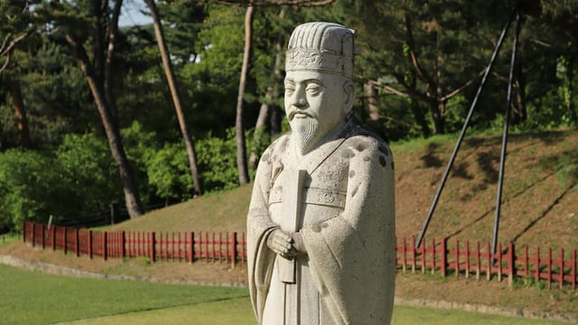 Stone statue of a historical figure in a green park with trees and grass on a sunny day.