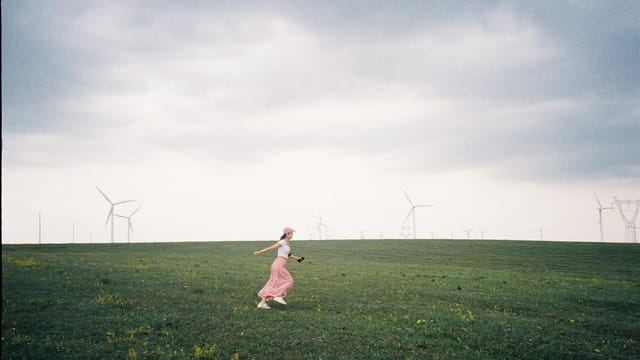 A woman runs joyfully in a spacious field with wind turbines on a cloudy day in Inner Mongolia.