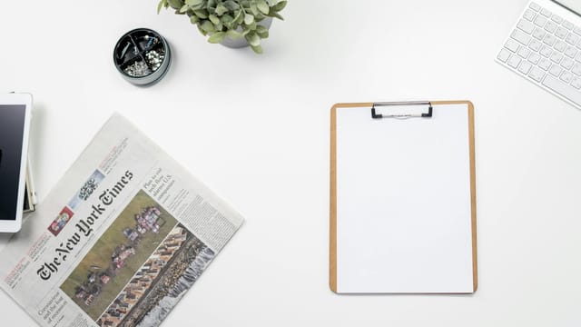Clean flatlay of a home office desk with The New York Times, clipboard, and keyboard.