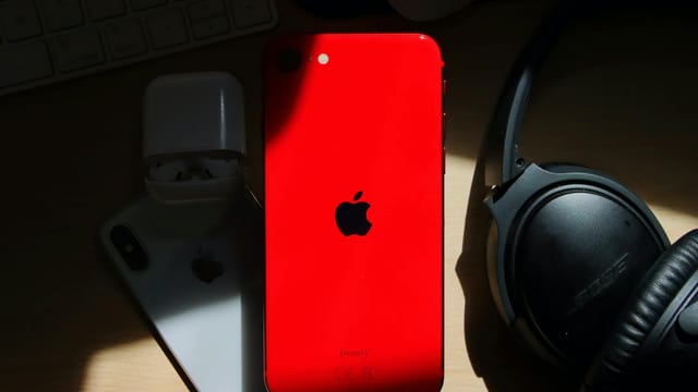 A flat lay of a red iPhone surrounded by AirPods and Bose headphones on a wooden surface.