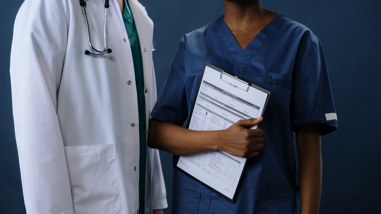 Two healthcare professionals in scrubs and lab coat with a clipboard, representing teamwork.