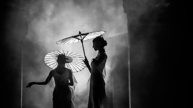 Black and white image of two women holding umbrellas in a mist, capturing elegance and Asian culture.
