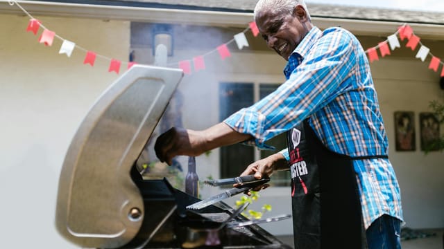Senior man barbecuing outside on a sunny day, enjoying cooking on a grill.