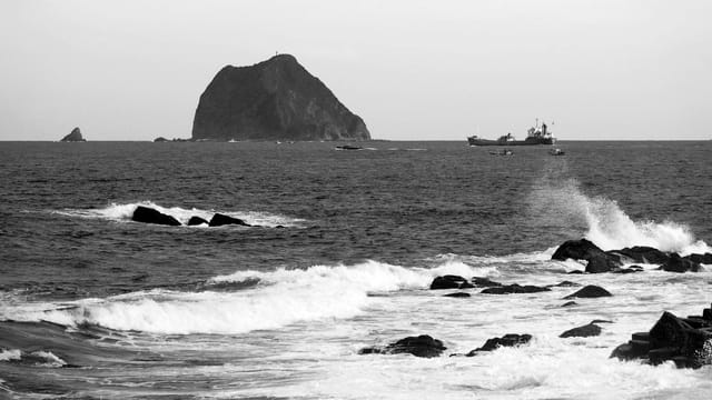 Black and white seascape featuring waves crashing on rocks, boats, and an island in Taiwan.
