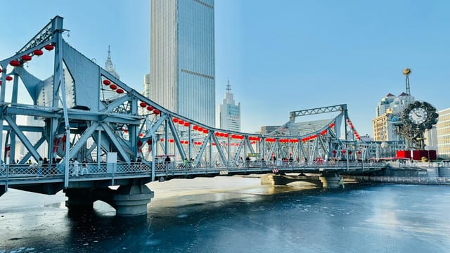 A scenic winter view of a bridge over a frozen river in Tianjin, China, with skyscrapers in the background.