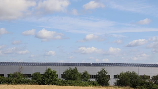 A countryside warehouse with solar panels and fields under a blue sky with clouds.