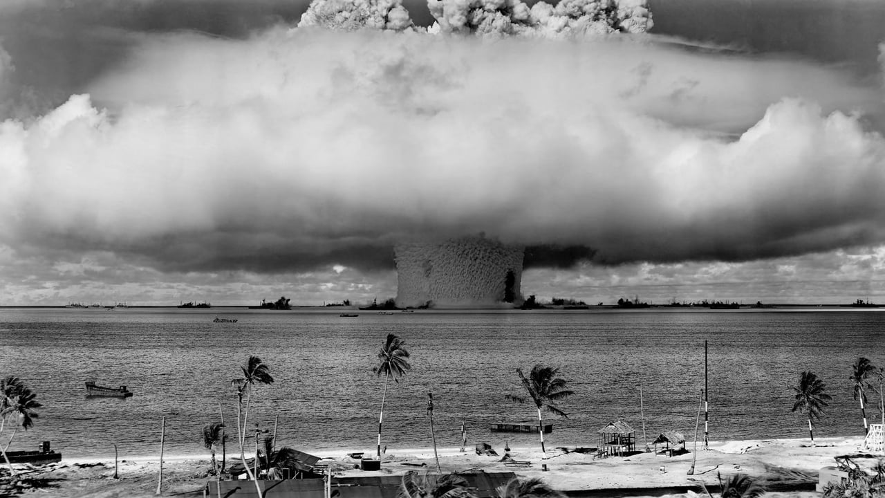 A black-and-white image of a nuclear explosion seen from a tropical beach with coconut trees.