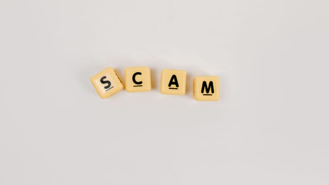 Four Scrabble tiles forming the word 'SCAM' on a neutral background, highlighting concepts like deceit.