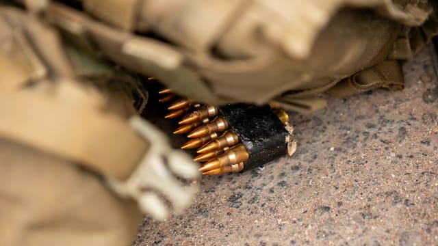 Detailed view of an ammunition belt partially covered by camouflage gear on a rough surface.