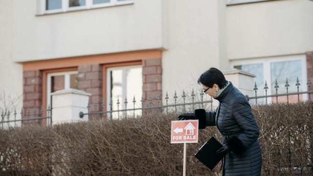 Real estate agent in a black coat placing a 'For Sale' sign in front of a house.