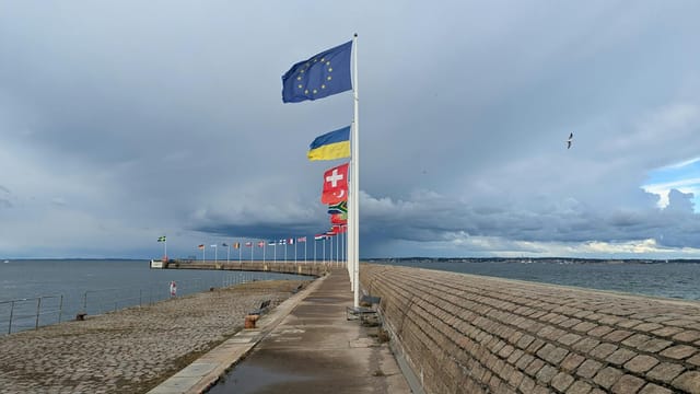 A pier adorned with European flags stretches into the sea under a dynamic cloudy sky.
