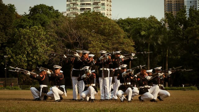 Uniformed soldiers performing a drill in a park with urban backdrop.