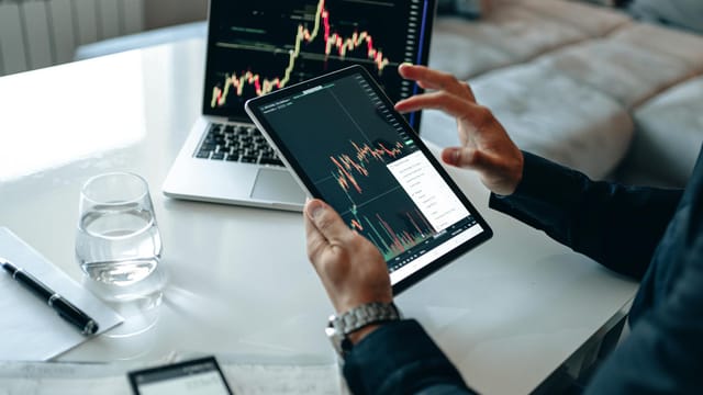 Close-up of a person using a tablet for online trading analysis with a laptop and glass of water nearby.