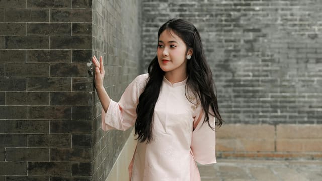 A woman in a pink traditional outfit poses thoughtfully against a textured brick wall.