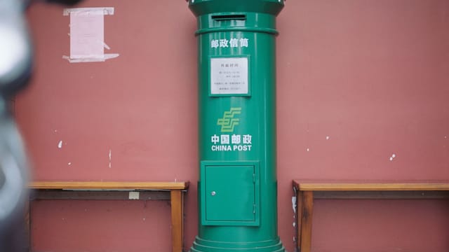 A China Post mailbox stands against a red wall, framed by benches, in a typical urban setting.