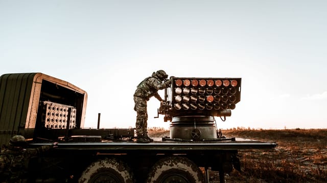 Soldier adjusting artillery on military truck in an open field under clear sky.