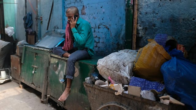 Man sitting in an urban slum using a smartphone amidst garbage and debris, New Delhi, India.