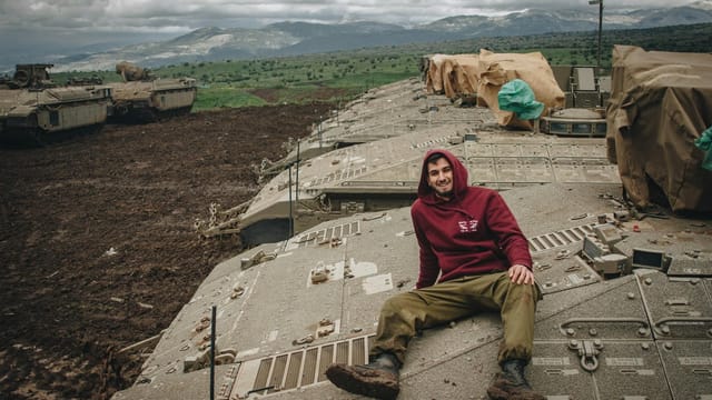 Young man in red hoodie sits on a tank in rural Lebanon, smiling.