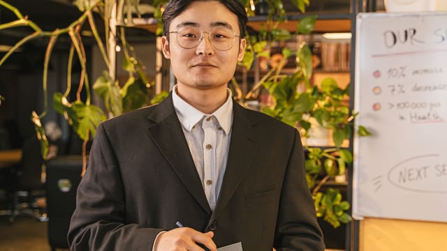 A confident young businessman in a suit and eyeglasses standing in a modern office.