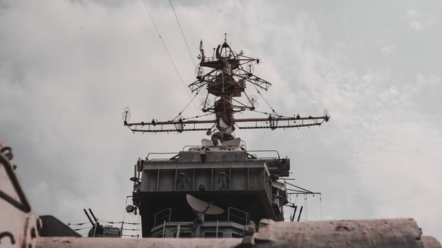 Close-up view of a military warship's radar and antenna mast against a cloudy sky.