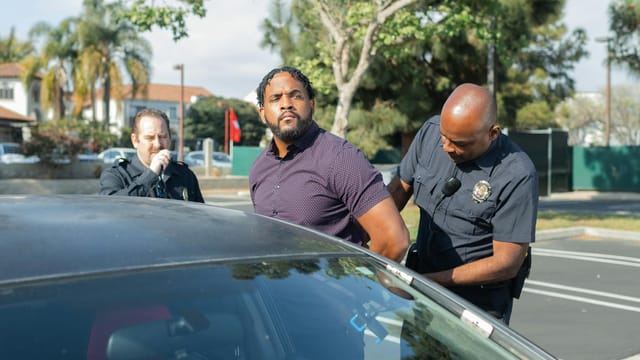 Two police officers detaining a suspect beside a car in a sunny outdoor setting.