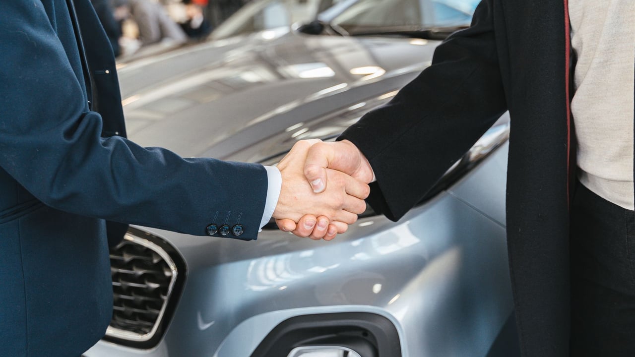 A close-up of two people shaking hands in front of a car in an indoor setting.