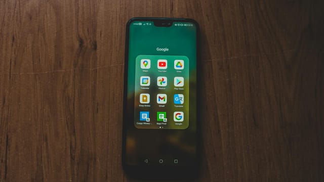 A smartphone showcasing various Google apps on a wooden table.